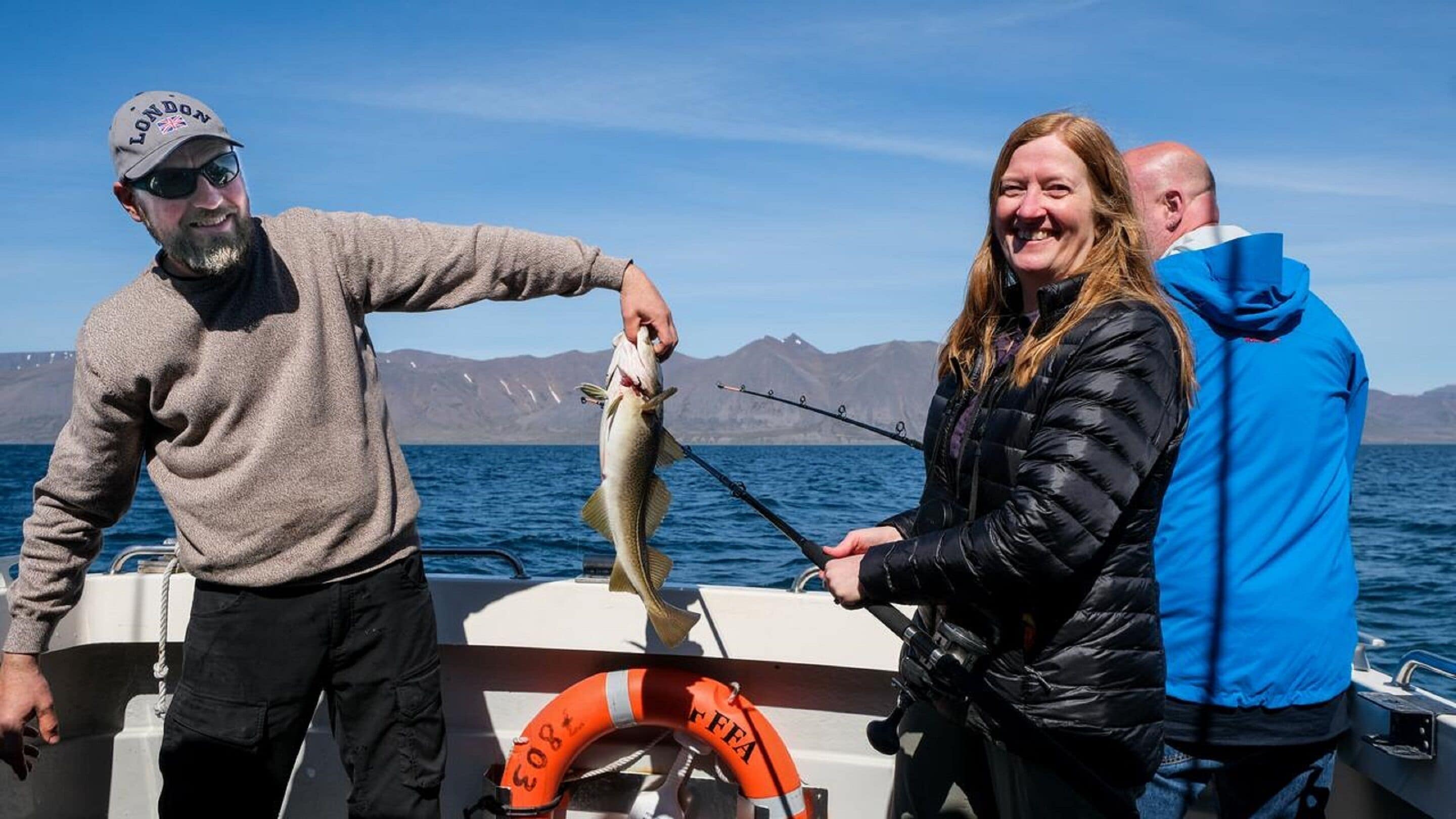 A boat with a person holding a fish, another person holding a fishing rod, and a life preserver on the deck.