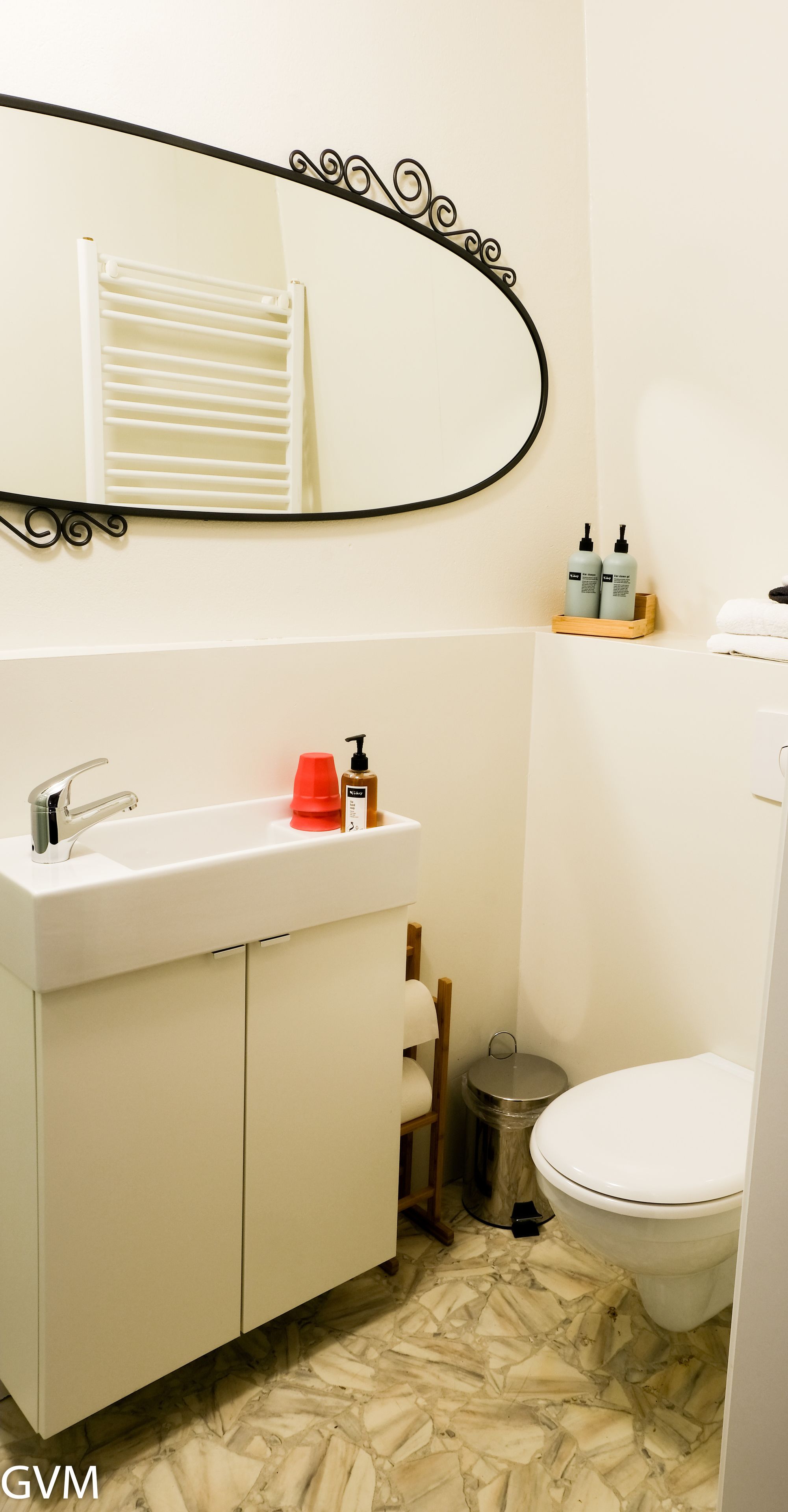 Une salle de bain avec un lavabo blanc, un miroir et des toilettes. On trouve des articles de toilette sur le lavabo et une poubelle à côté des toilettes.