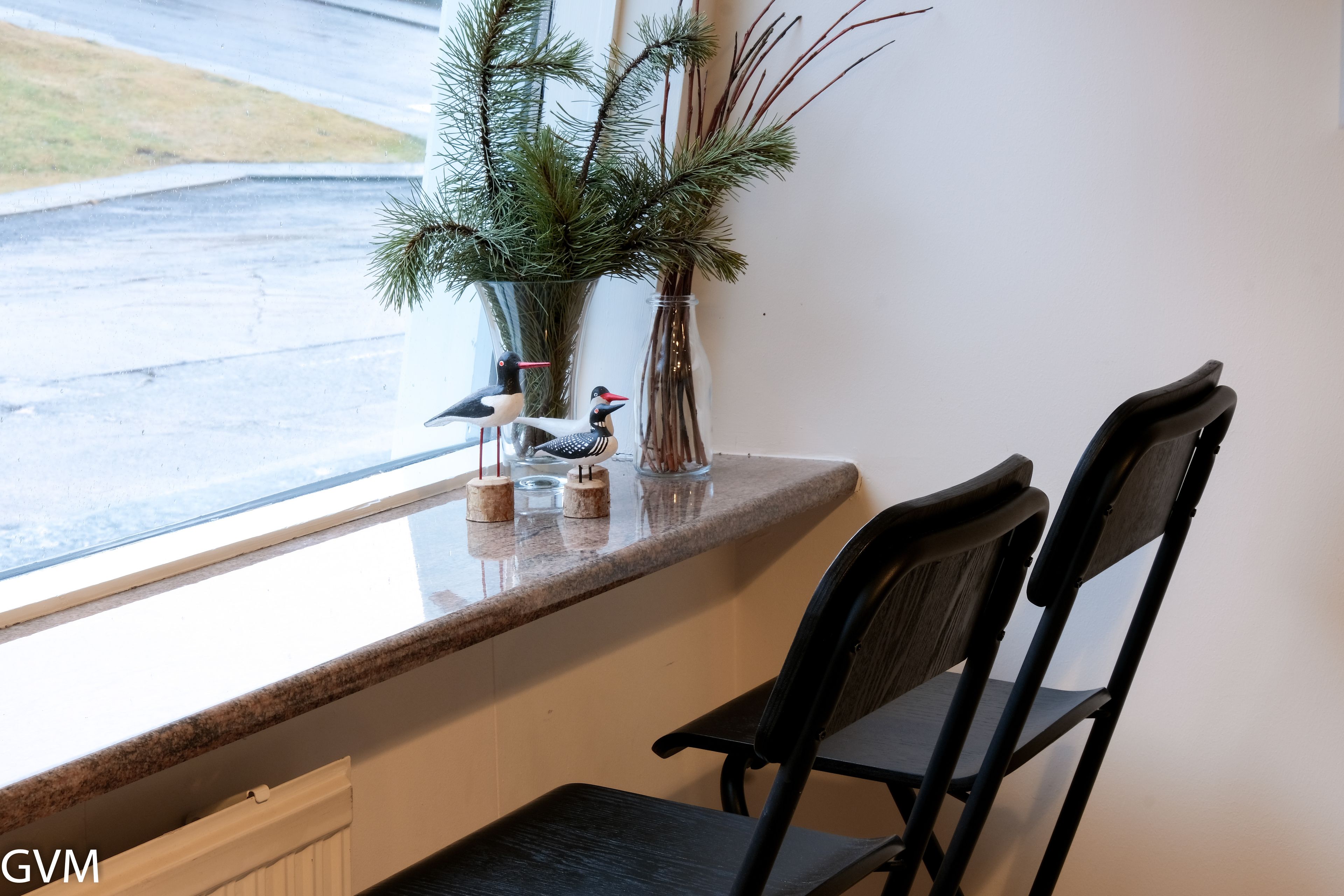 Un banc de fenêtre avec deux chaises, un vase décoré de branches de pin et une petite figurine d’oiseau.