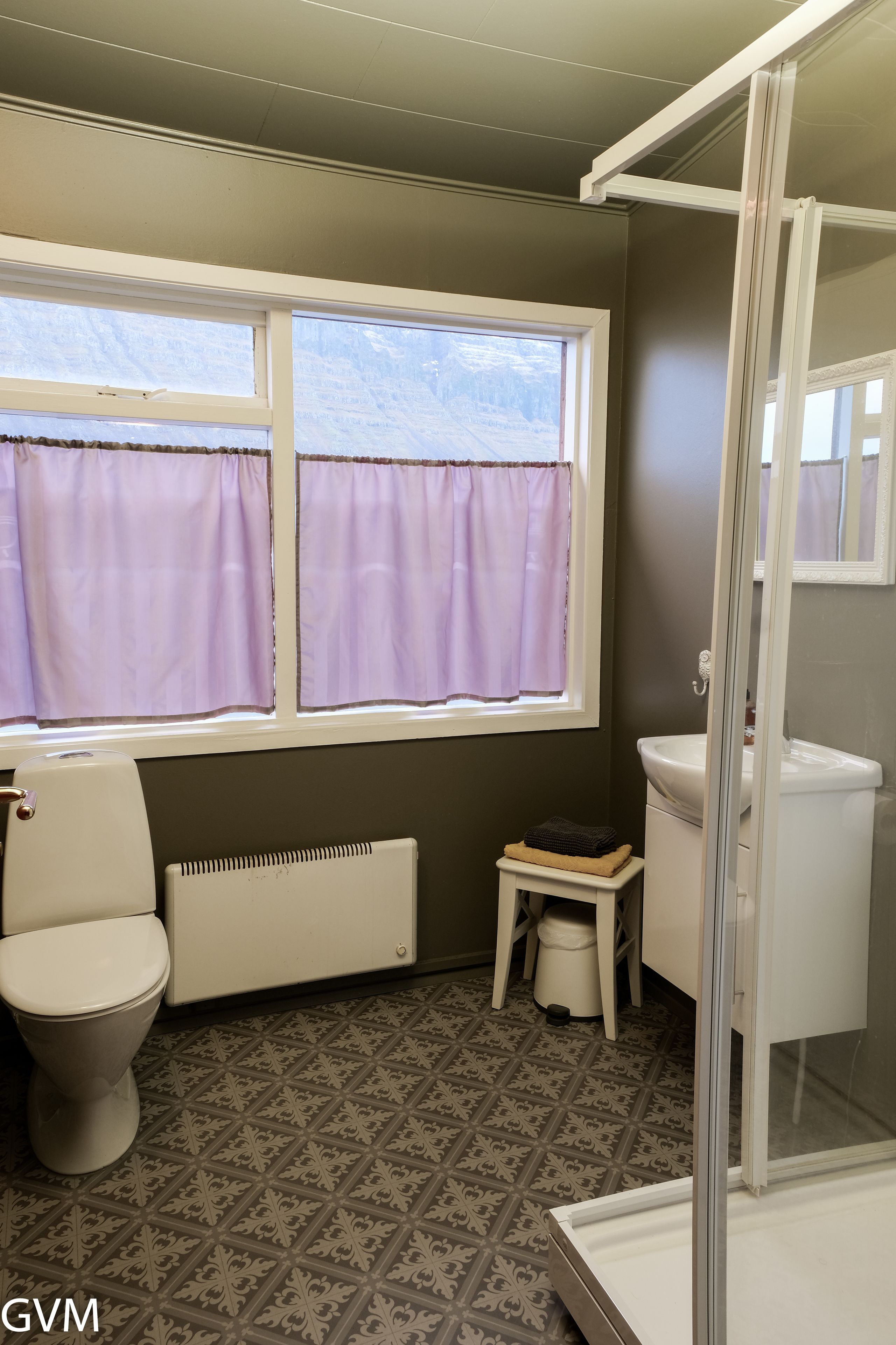 A bathroom with a toilet, a radiator, a small stool, a sink, and a window with purple curtains.