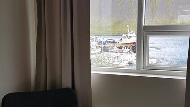 A room with a bed, a chair, and a window with a view of boats and a hilly landscape. - Harbour Inn guesthouse (Vesturbyggð)