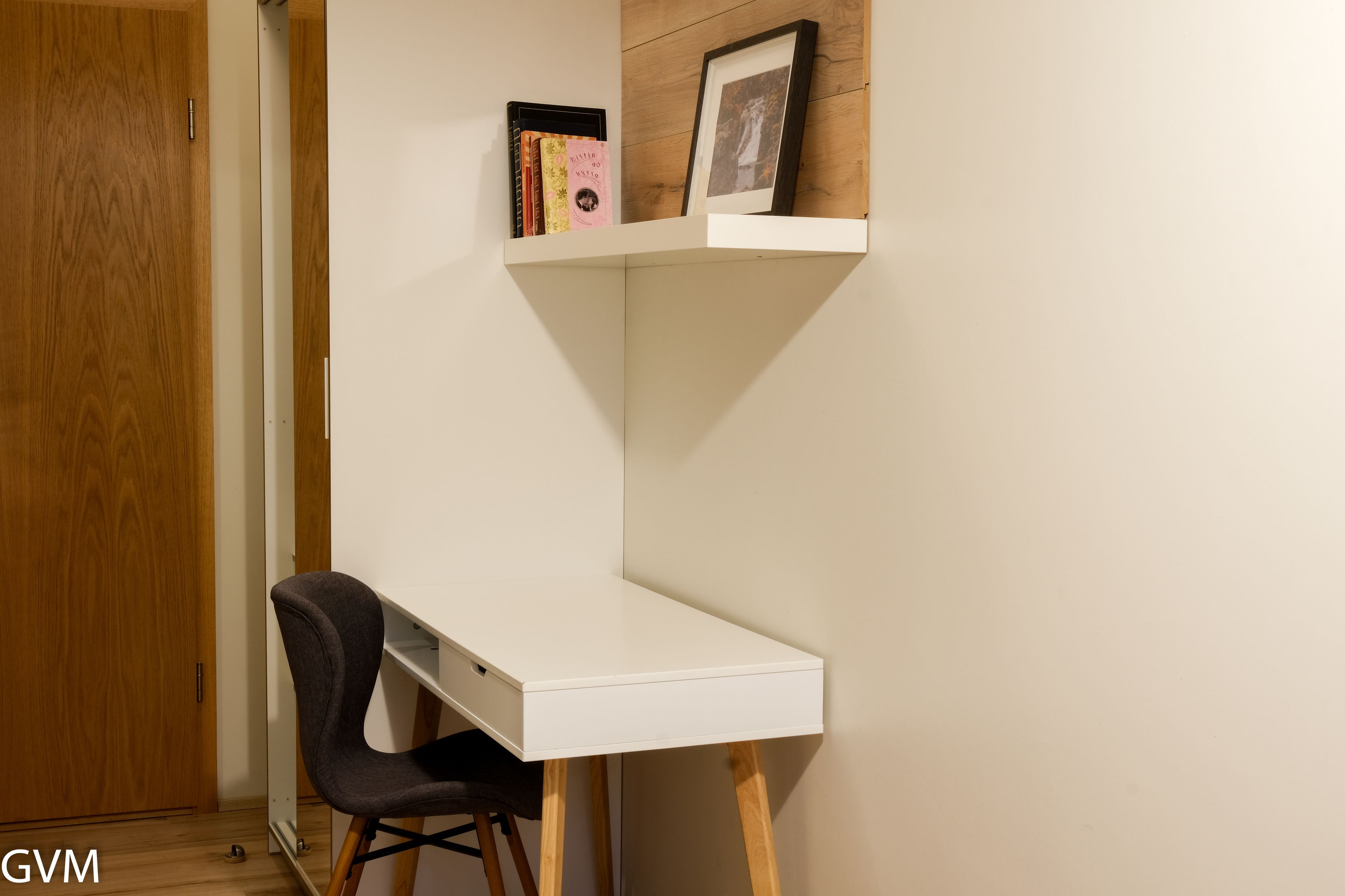 Un escritorio en esquina con una silla, una puerta de madera, y una estantería con libros y un marco de fotos.