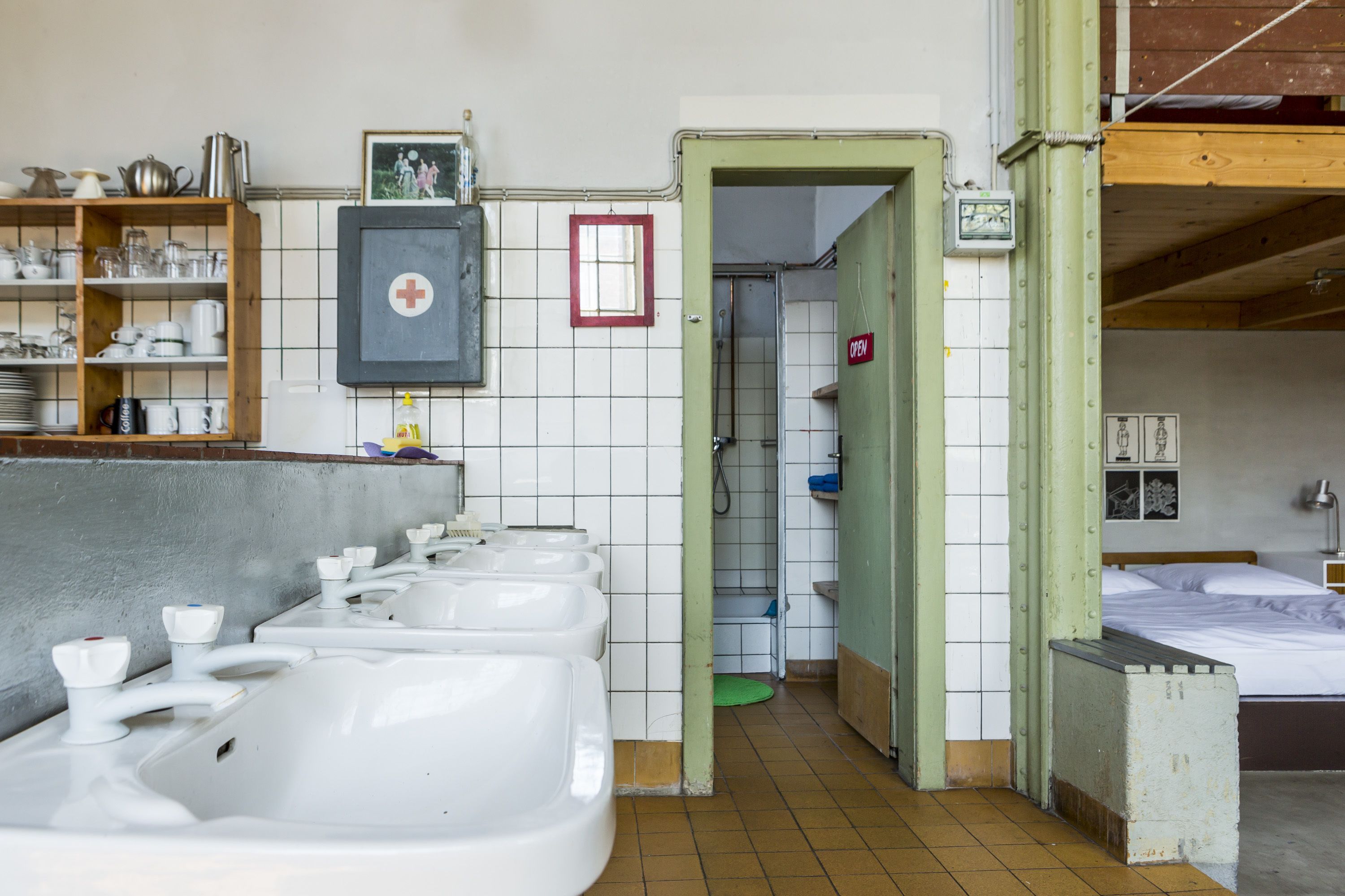 Baño público con varios lavamanos, una ducha y una cama en la habitación contigua.