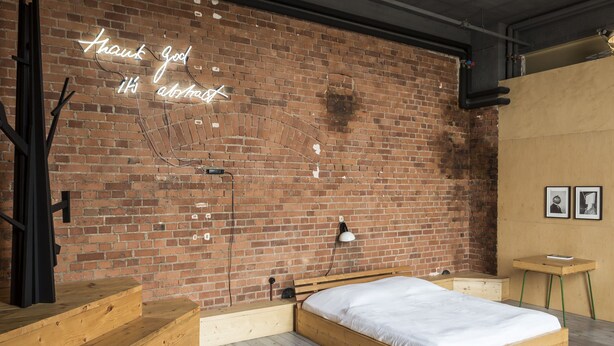 Una stanza con parete in mattoni, un letto, una panca in legno, un tavolino e un neon.