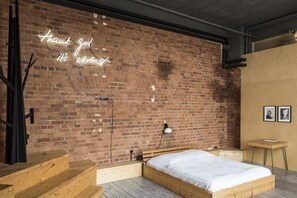 Una habitación con pared de ladrillo, una cama, un banco de madera, una mesita y un letrero de neón.