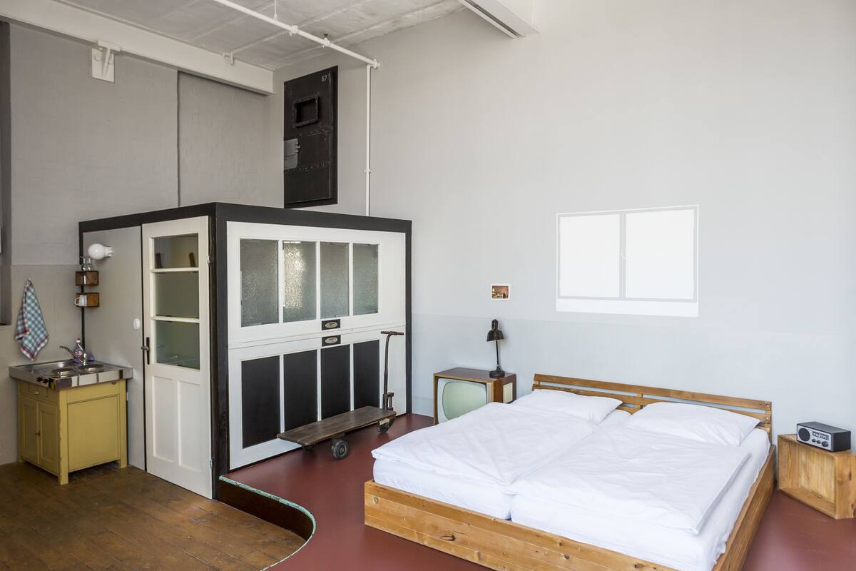 A room with a bed, a small kitchenette, a window, and a ceiling-mounted storage unit.