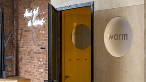 Una porta gialla con una finestra circolare, un'insegna con la scritta 'warm' sul muro, e un muro di mattoni con un neon.