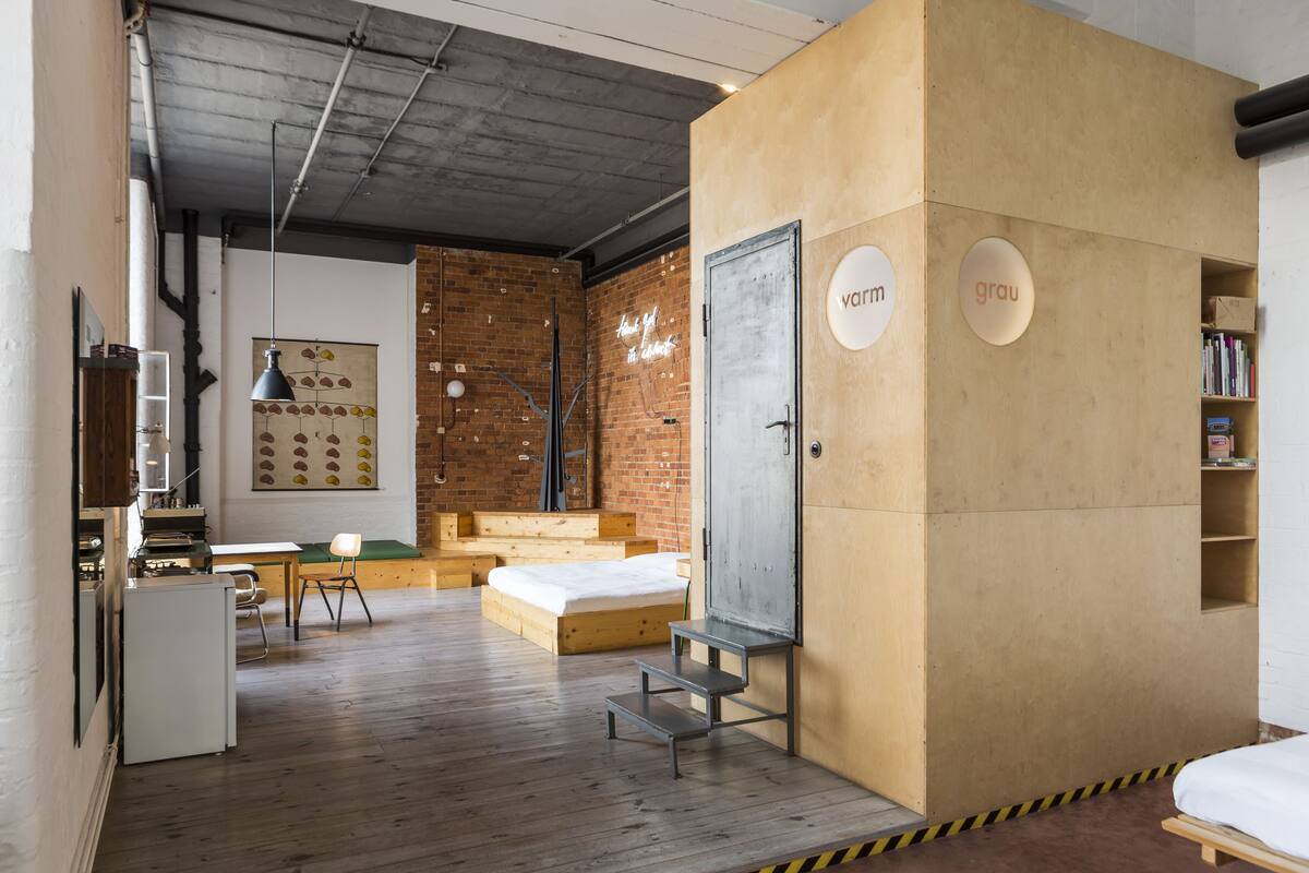 A modern, compact room with a wooden floor, a bed, a desk, and a chair. There is a bookshelf with books and a neon sign on the wall.