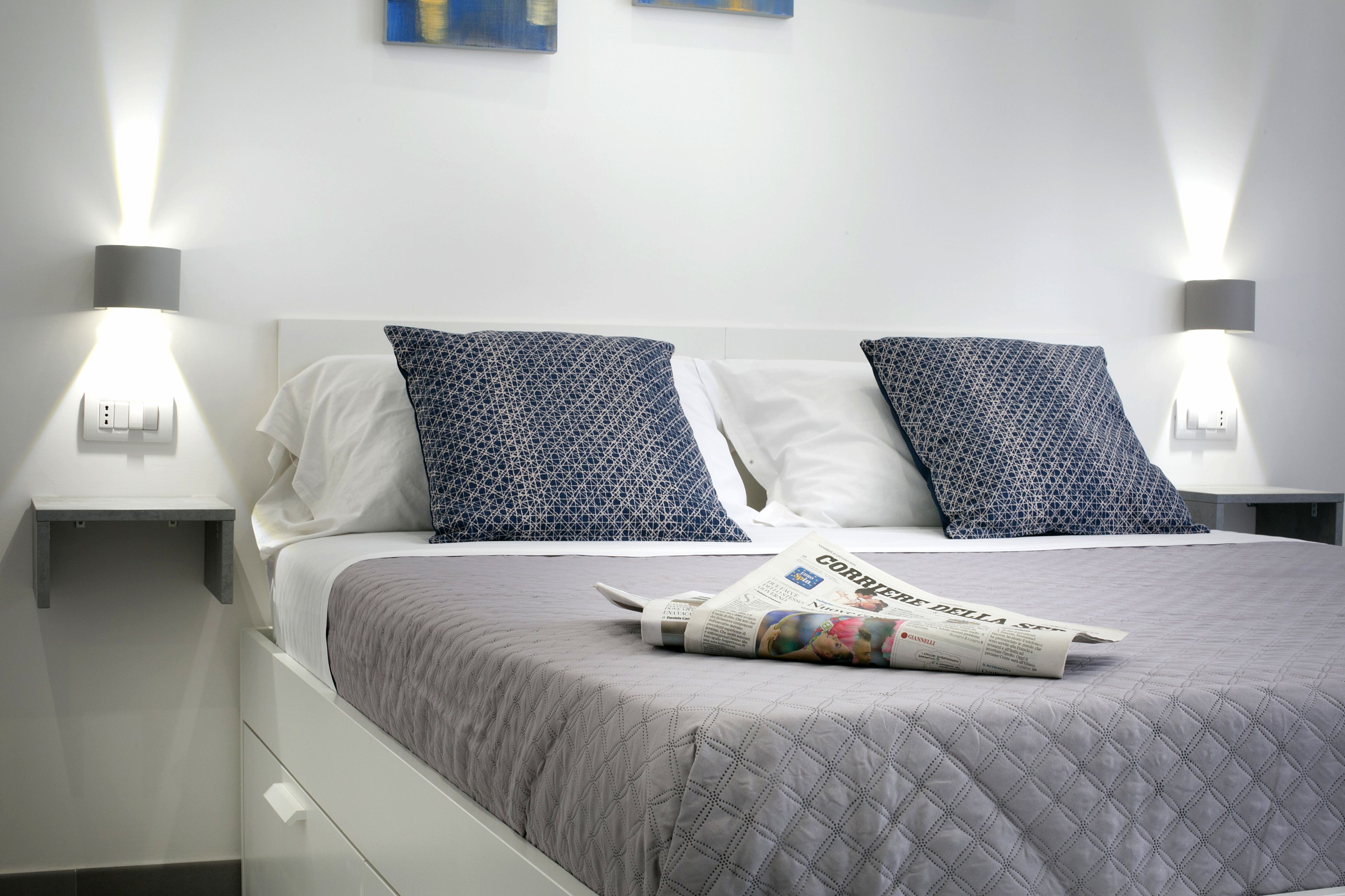 A neatly made bed with a quilted grey bedspread, two pillows with a geometric pattern, and a folded newspaper on the bed.