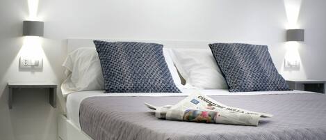 A neatly made bed with a quilted grey bedspread, two pillows with a geometric pattern, and a folded newspaper on the bed.