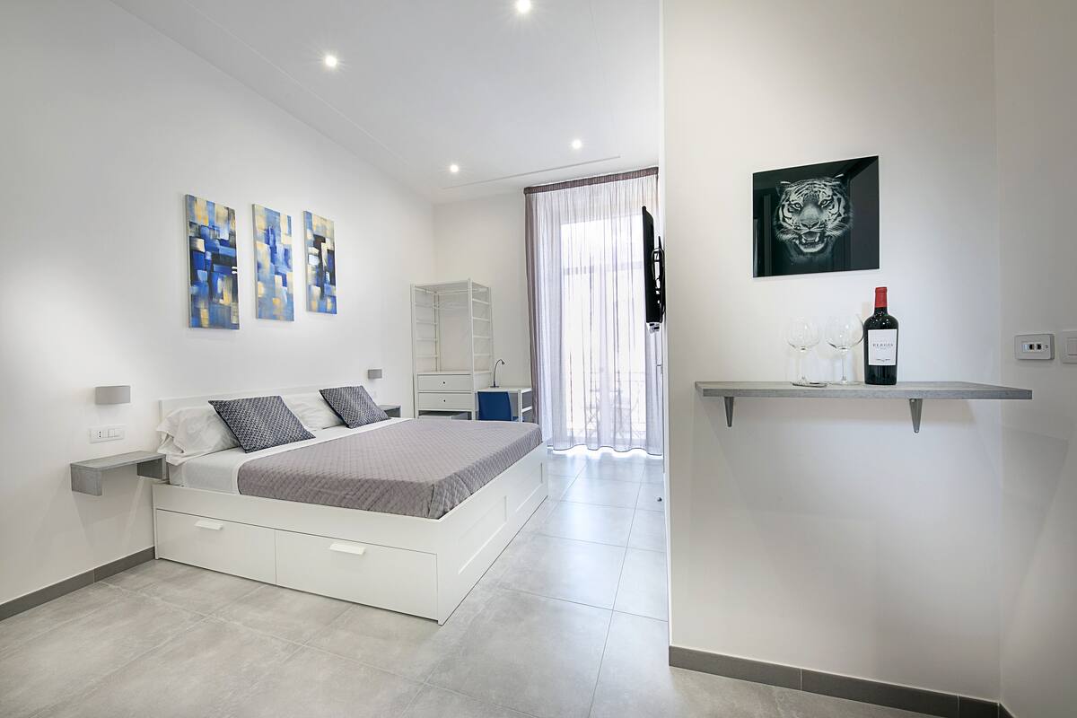 A modern bedroom with a bed, a wardrobe, a shelf with a bottle and glasses, and abstract art on the wall.