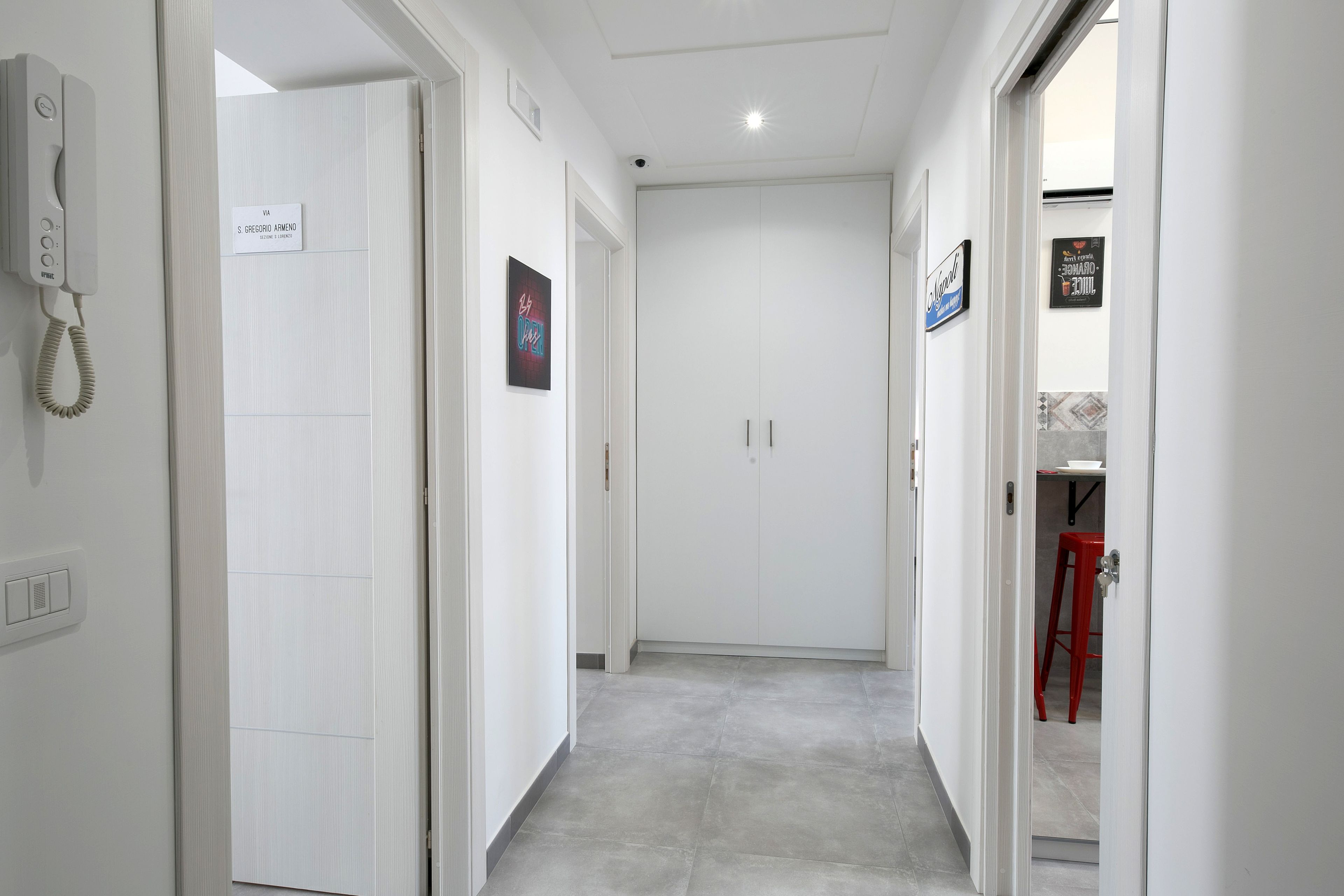 A hallway with a phone on the left, a sign reading "VA S. GREGORIO AMENDO" on the right, and a red stool in the background.