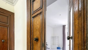 A hallway with a wooden door leading to a bedroom with a bed, a dresser, and a chair.