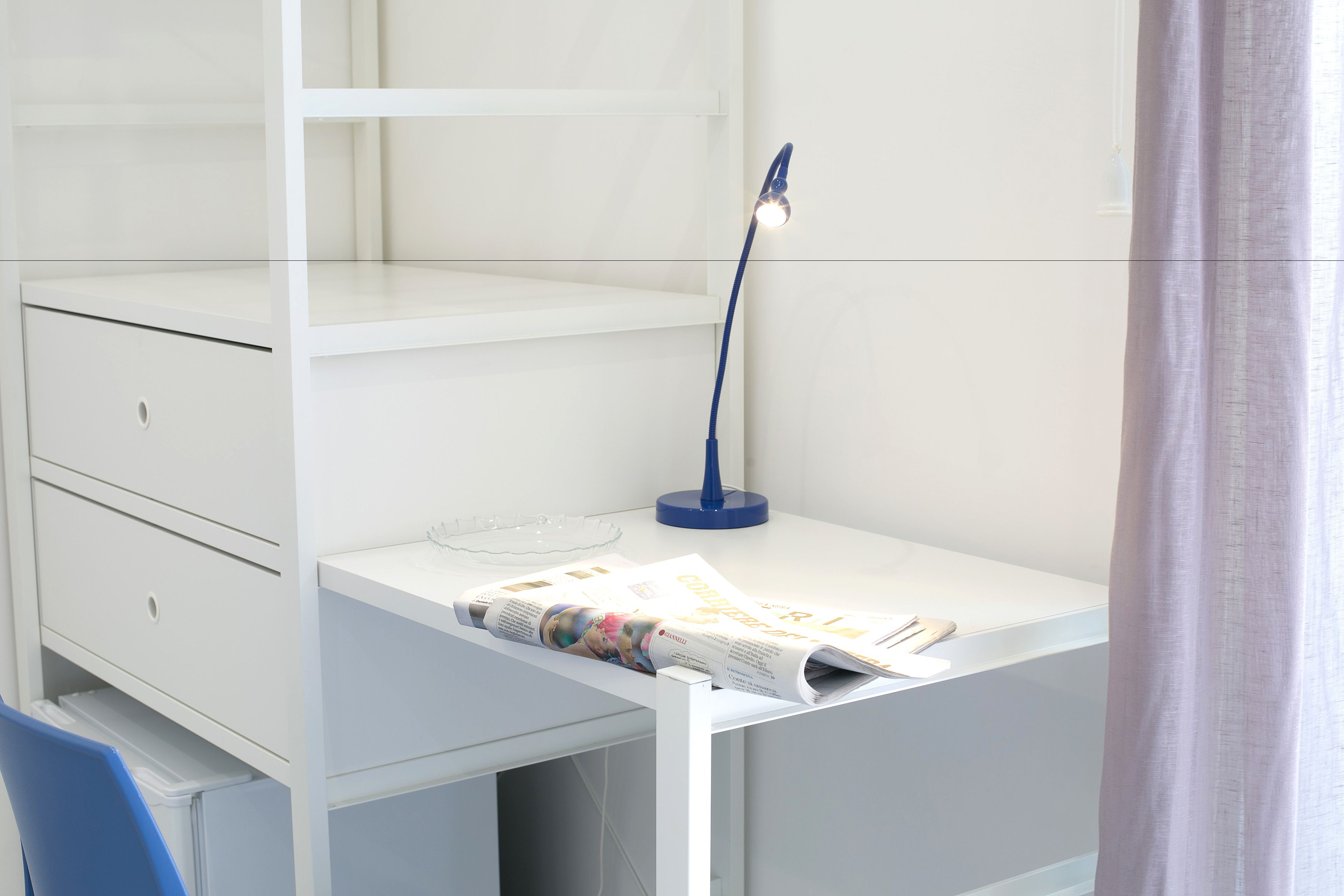 A white desk with a magazine, a blue desk lamp, and a blue chair.