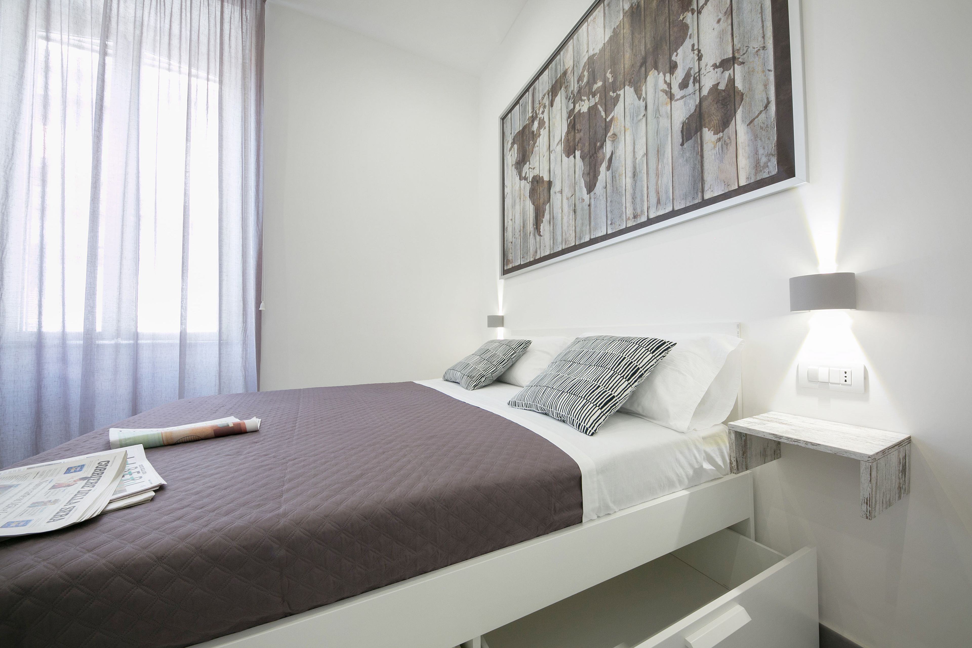 Una cama bien hecha con cubrecama marrón y ropa de cama blanca, un cuadro de un mapa mundial en la pared, una mesita de noche con una lámpara y una ventana con cortinas de encaje.