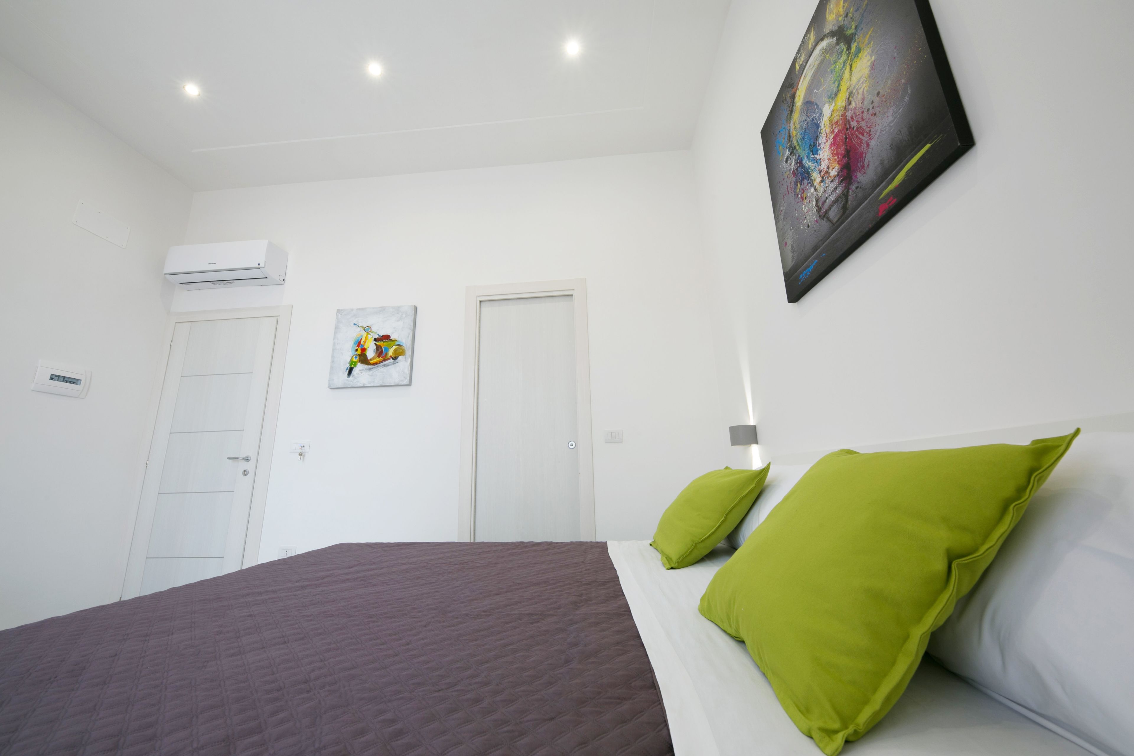 Un dormitorio con cama, almohadas verdes, una puerta blanca y un cuadro abstracto colorido en la pared.