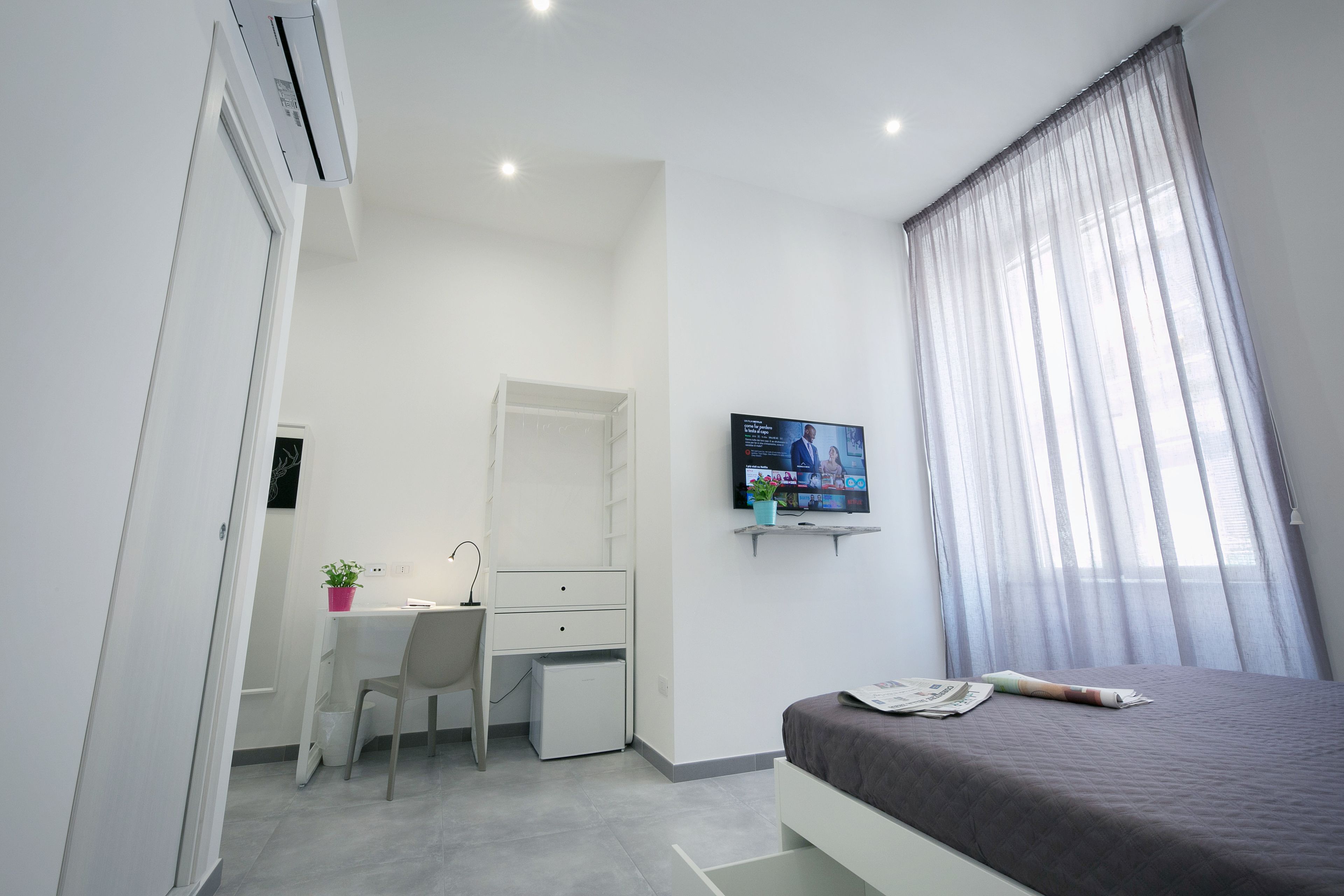 A modern bedroom with a bed, a desk, a chair, a television, and a window with curtains.