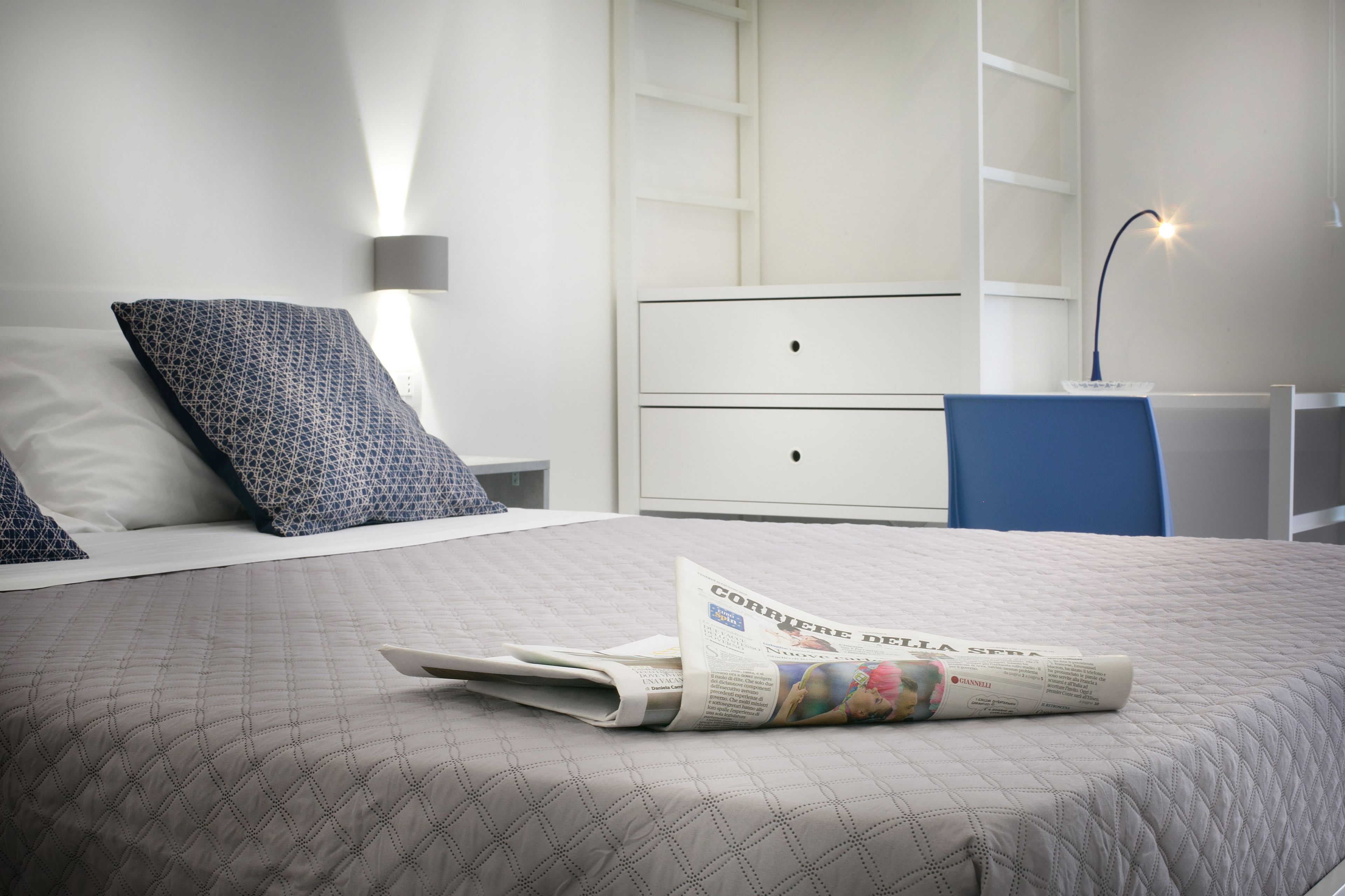 Una cama bien hecha con una colcha estampada, un periódico doblado y una silla azul en un dormitorio moderno con muebles blancos y una lámpara de noche.