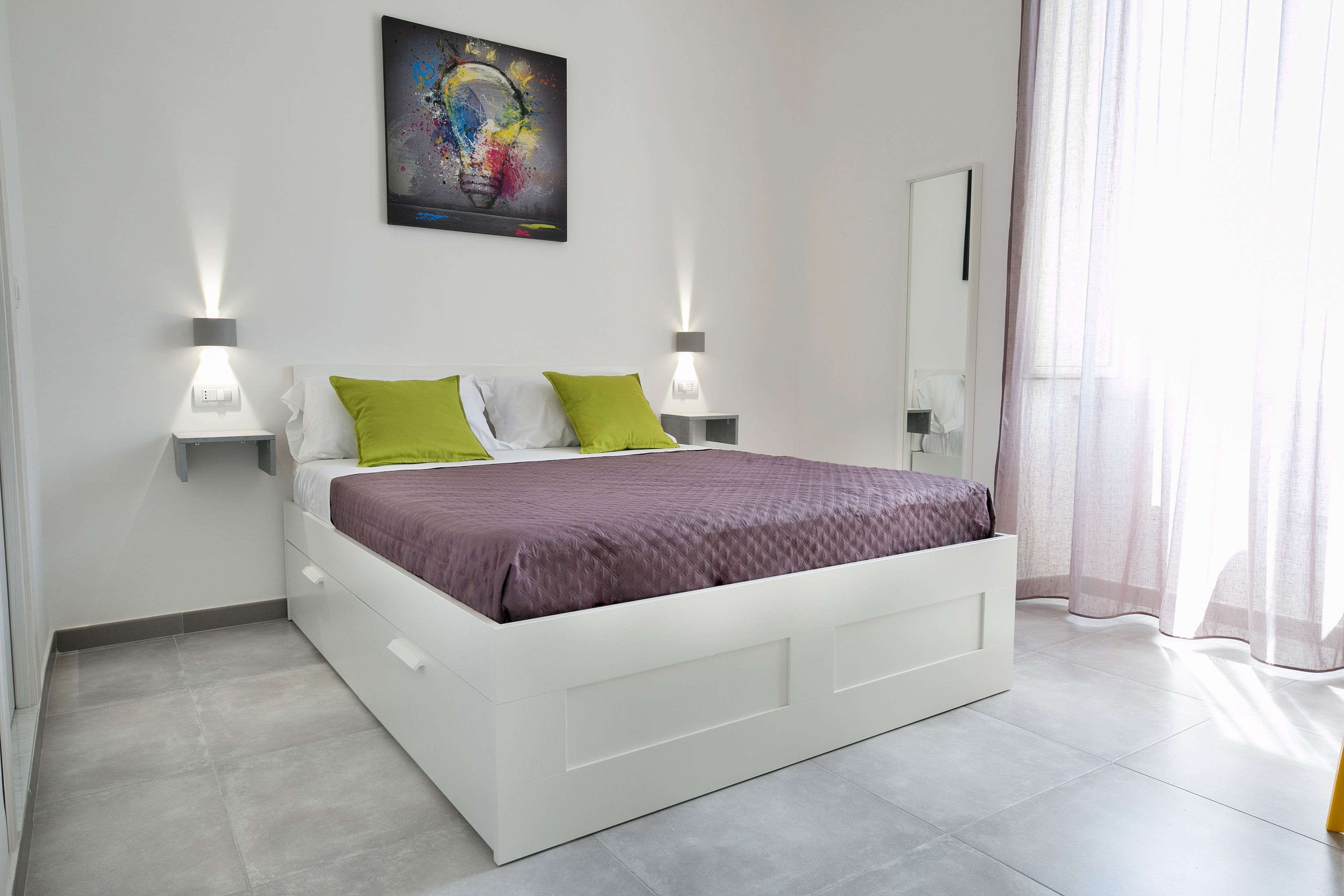 Une chambre moderne avec un lit blanc, des coussins verts et une couvre-lit violette. On y trouve deux lampes de chevet et un grand miroir accroché au mur.