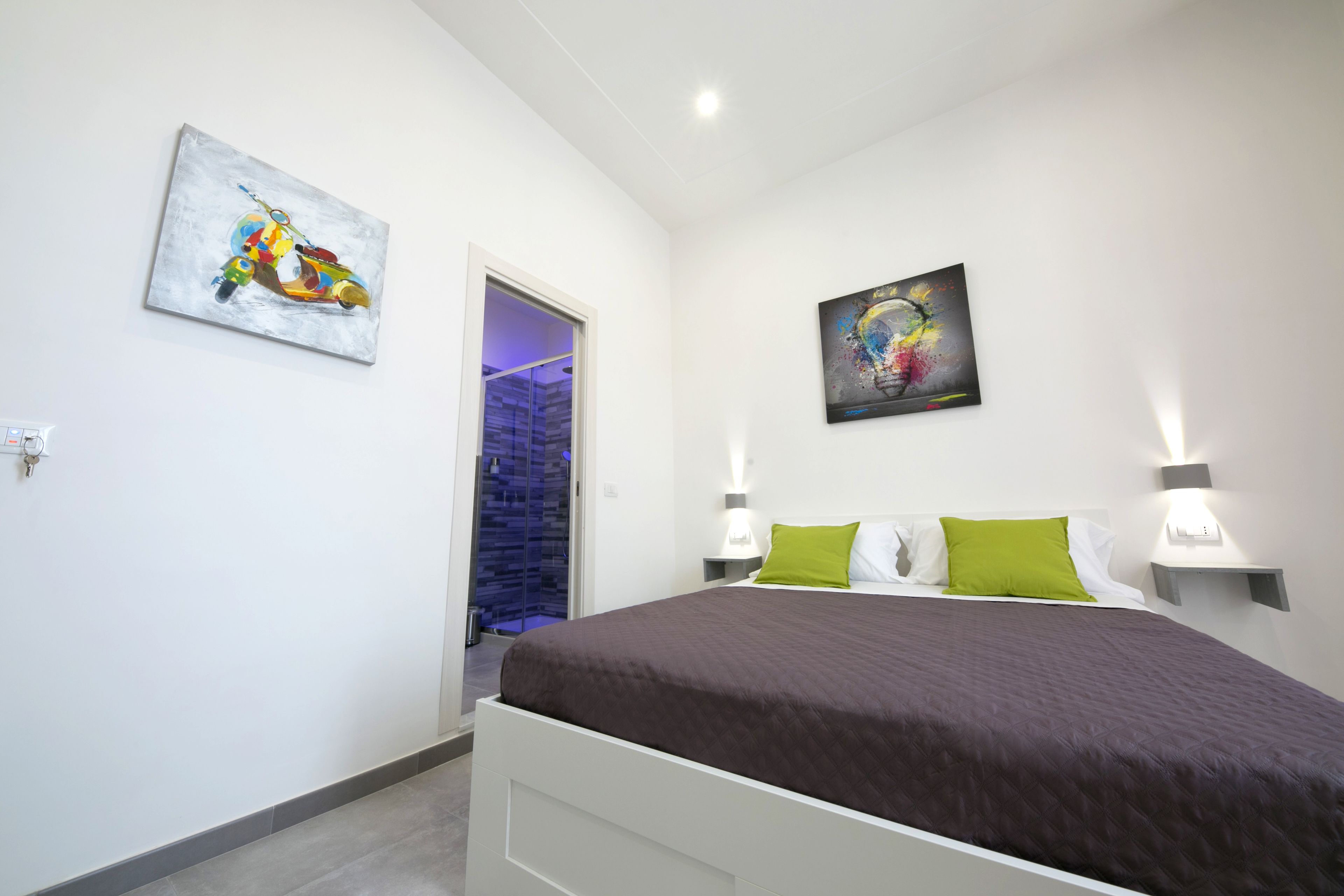 Une chambre à coucher comprenant un lit, deux lampes fixées au mur, une porte donnant sur une salle de bain et deux tableaux accrochés aux murs.