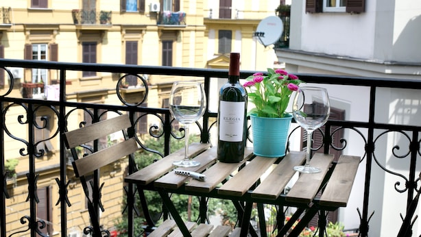 A balcony with a wooden table, a bottle of wine, two wine glasses, and a potted plant with pink flowers.