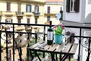 A balcony with a wooden table, a bottle of wine, two wine glasses, and a potted plant with pink flowers.