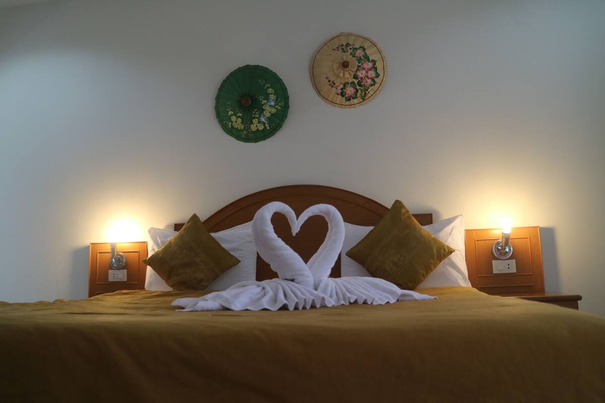 A neatly made bed with a heart-shaped towel arrangement, two wall-mounted lamps, and two decorative wall hangings.