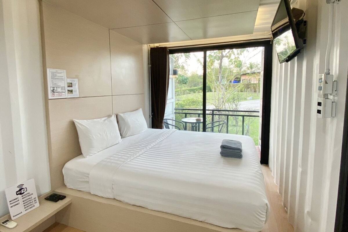 A modern hotel room with a large bed, a flat-screen TV mounted on the wall, and a view of the outdoors through a large window.