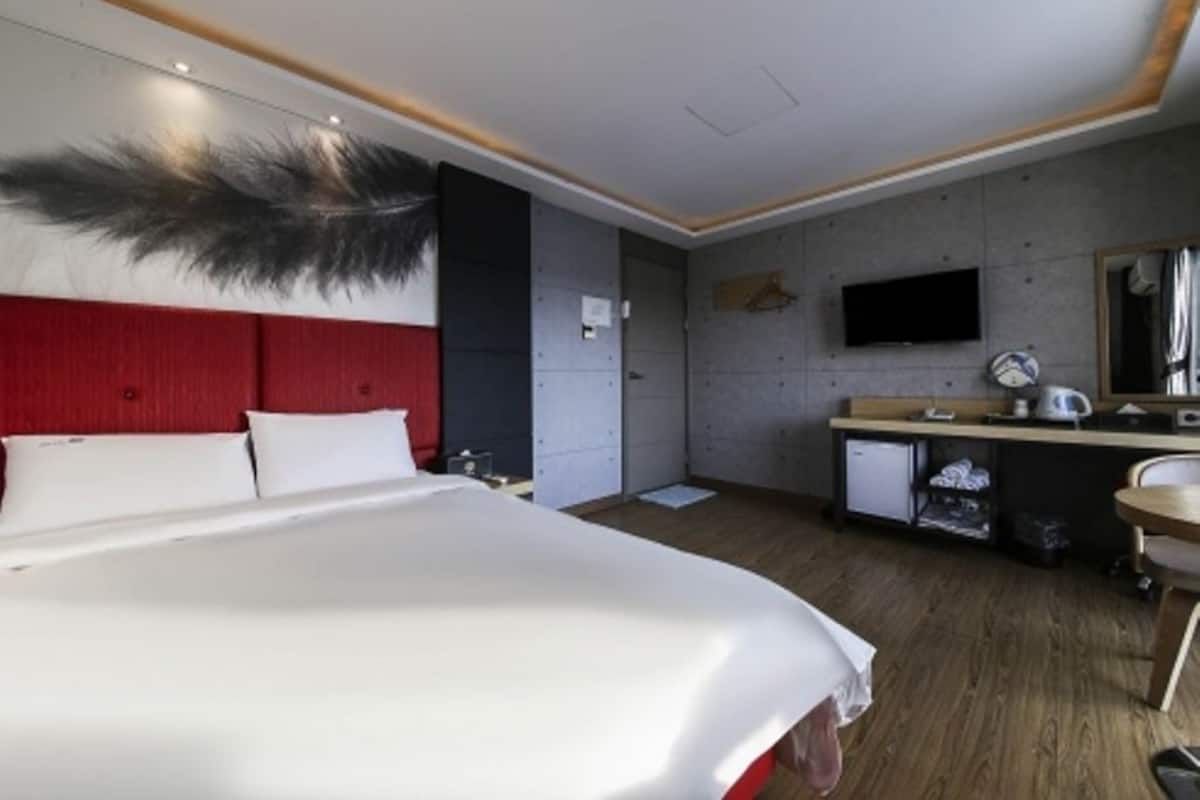 A hotel room with a large bed, a red headboard, a flat-screen TV mounted on the wall, a small kitchenette, and a wooden floor.