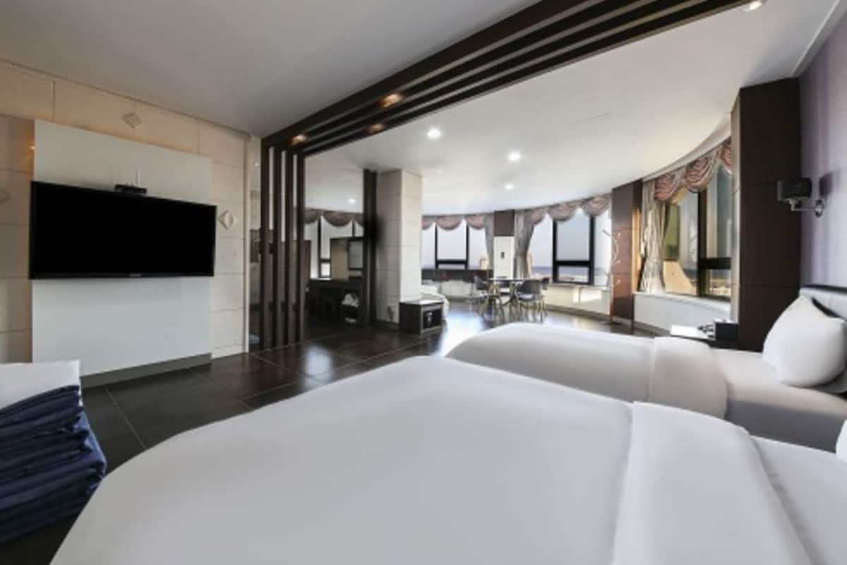 A modern hotel room with a large bed, a flat-screen TV, a dining area, and a view of the city.