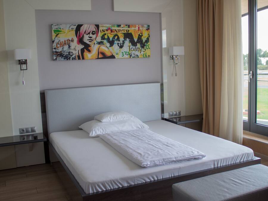 A modern hotel room with a large bed, a window with a view of the outdoors, and a colorful artwork on the wall.