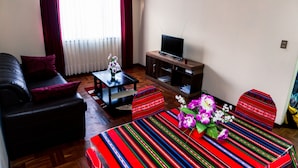 Ein Wohnzimmer mit einer Couch, einem Couchtisch, einem Fernseher auf einem Schrank und einer bunten, gestreiften Tischdecke.