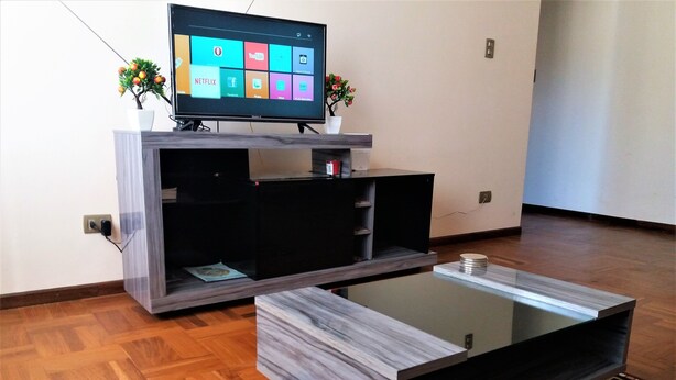 Ein modernes Wohnzimmer mit einem Flachbildfernseher auf einem Holzständer, einem Glastisch und einem gemusterten Teppich.