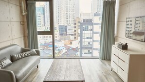 Un salon moderne avec un canapé gris, une table basse en bois et une commode blanche. De grandes fenêtres offrent une vue sur le paysage urbain.