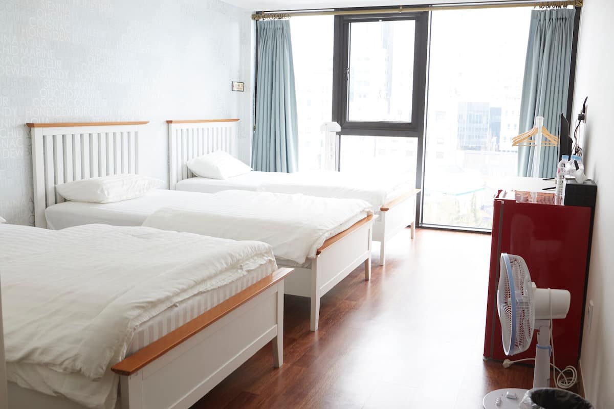 Une chambre d’hôtel avec deux lits simples, une commode rouge, un ventilateur sur pied et une grande fenêtre avec des rideaux.