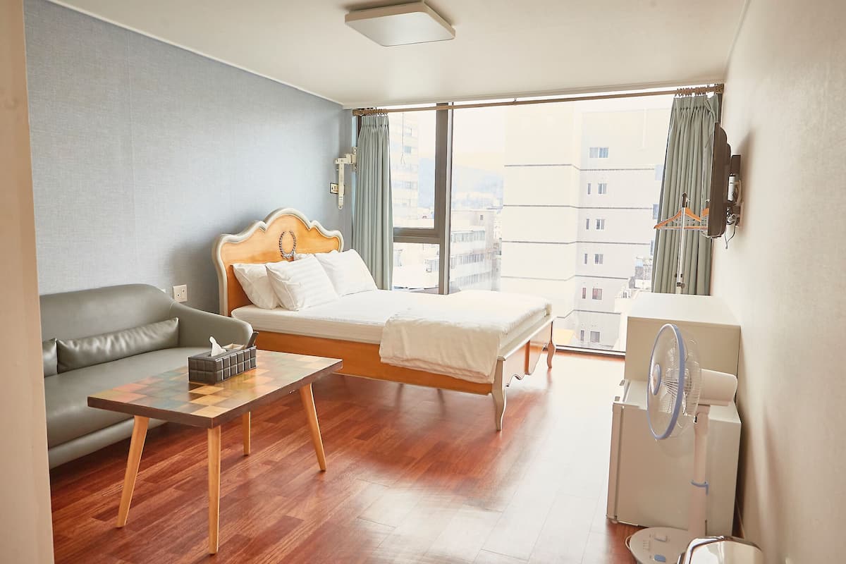 Une chambre d’hôtel avec un lit, un canapé, une petite table, un ventilateur sur pied et une fenêtre avec des rideaux.