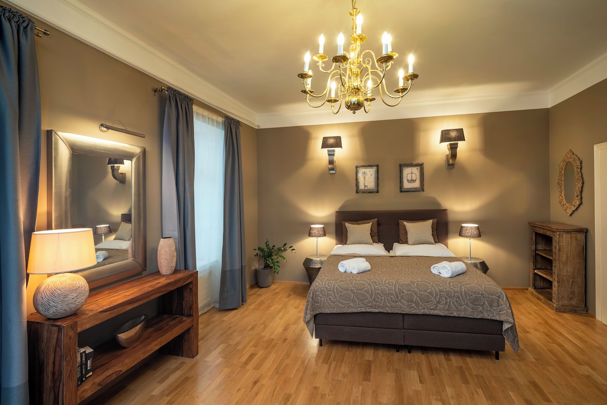 A bedroom with a large bed, bedside lamps, a mirror, a wooden console table, and a chandelier.