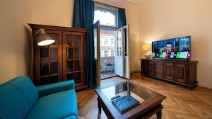 Ein Wohnzimmer mit einem blauen Sofa, einem Holztisch mit einem Laptop, einem Fernseher, der einen Nachrichtensender zeigt, und Blick auf ein Gebäude durch eine offene Tür.