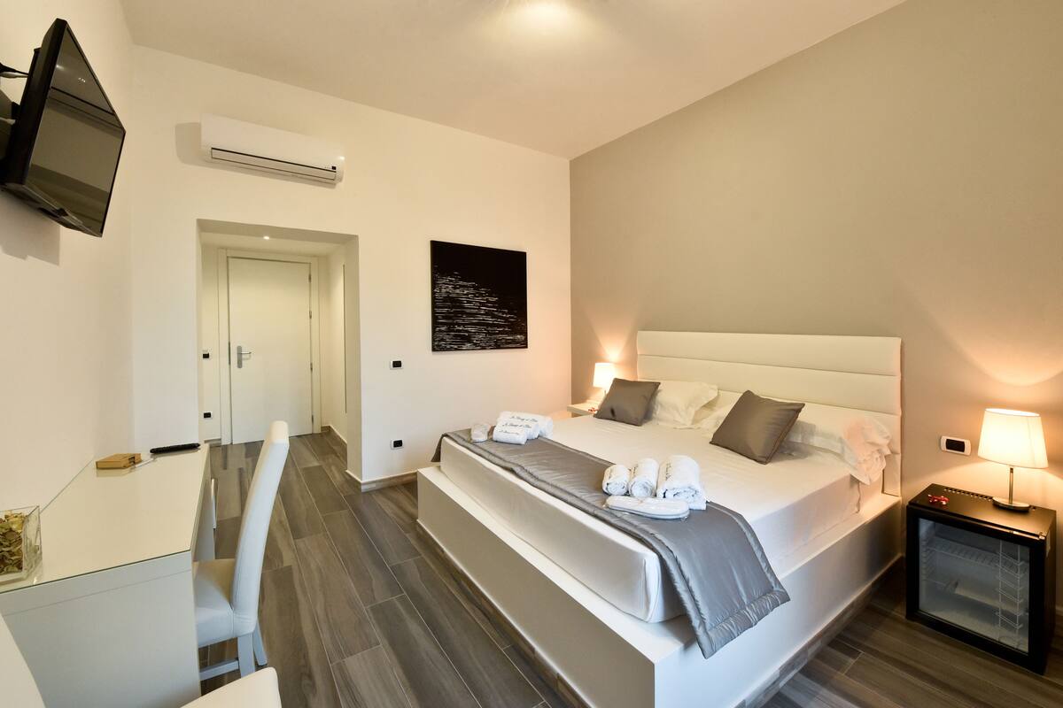 A modern hotel room with a large bed, bedside table, TV, air conditioning unit, and a small desk with chair.