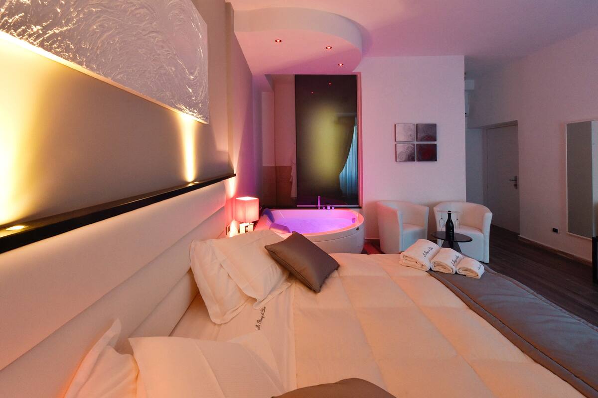 A modern hotel room with a large bed, a sitting area with a chair and a table, and a spa area with a hot tub.