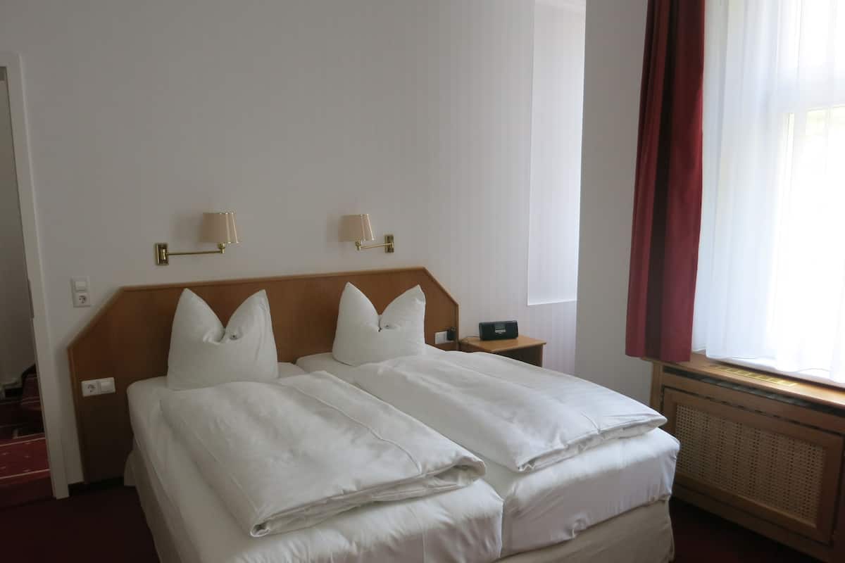 A hotel room with a double bed, two bedside lamps, a heater, and a window with curtains.
