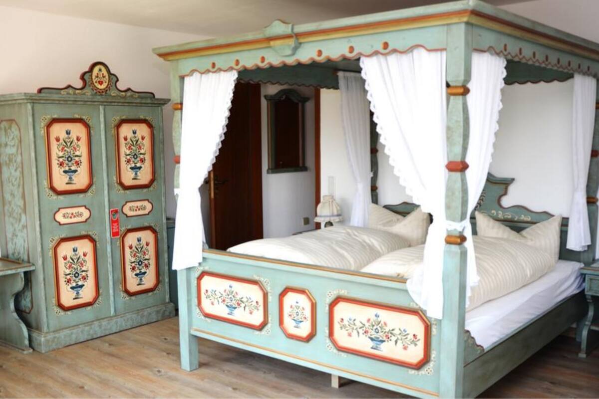 Ein Bett mit Baldachin, eine Kommode im Vintage-Stil mit floralen Verzierungen und ein kleiner Nachttisch.
