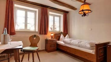 A bedroom with a wooden bed, a nightstand, a chair, a table with a lamp, and two windows with curtains.