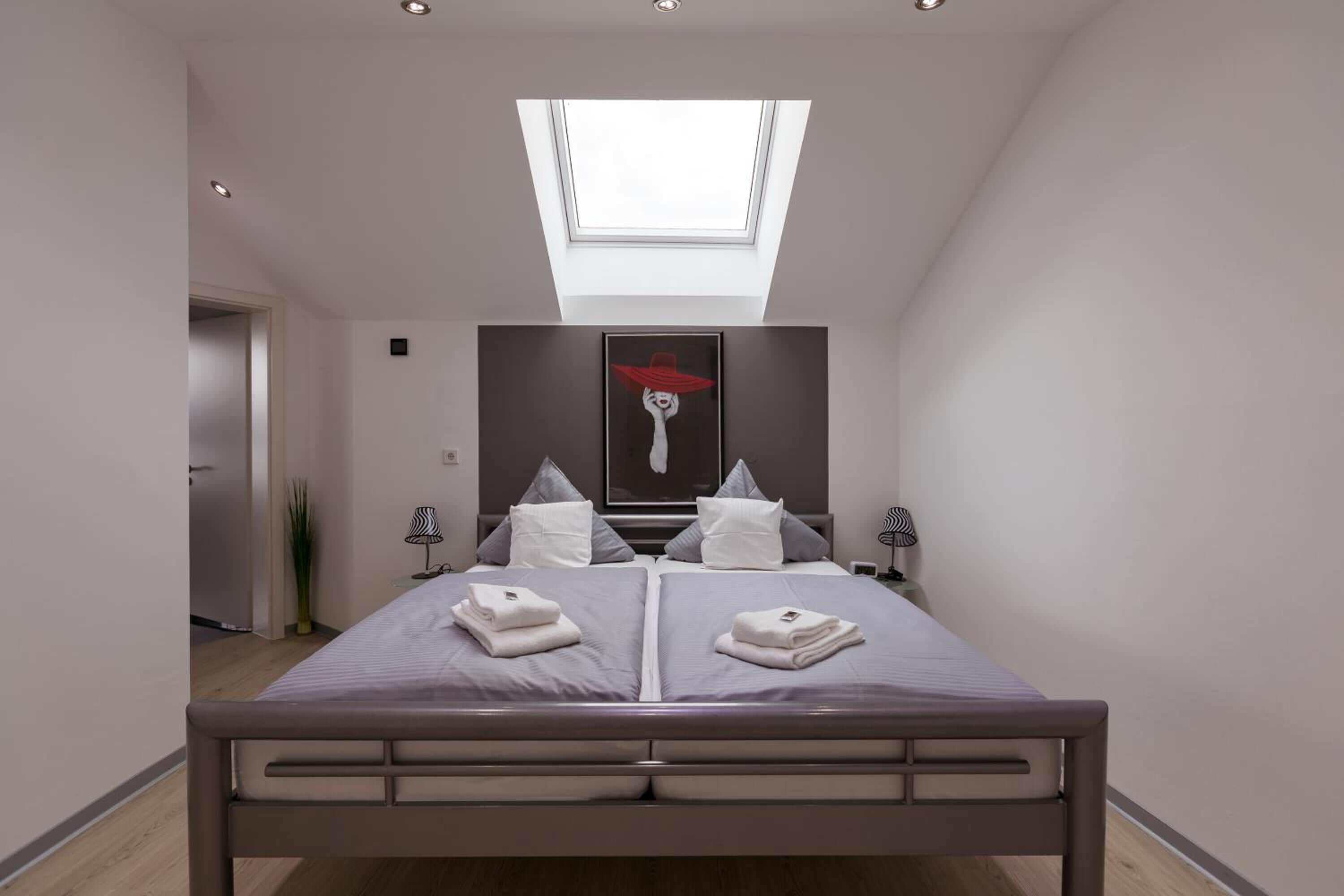 A modern bedroom with a skylight, a bed with pillows, and a painting on the wall.