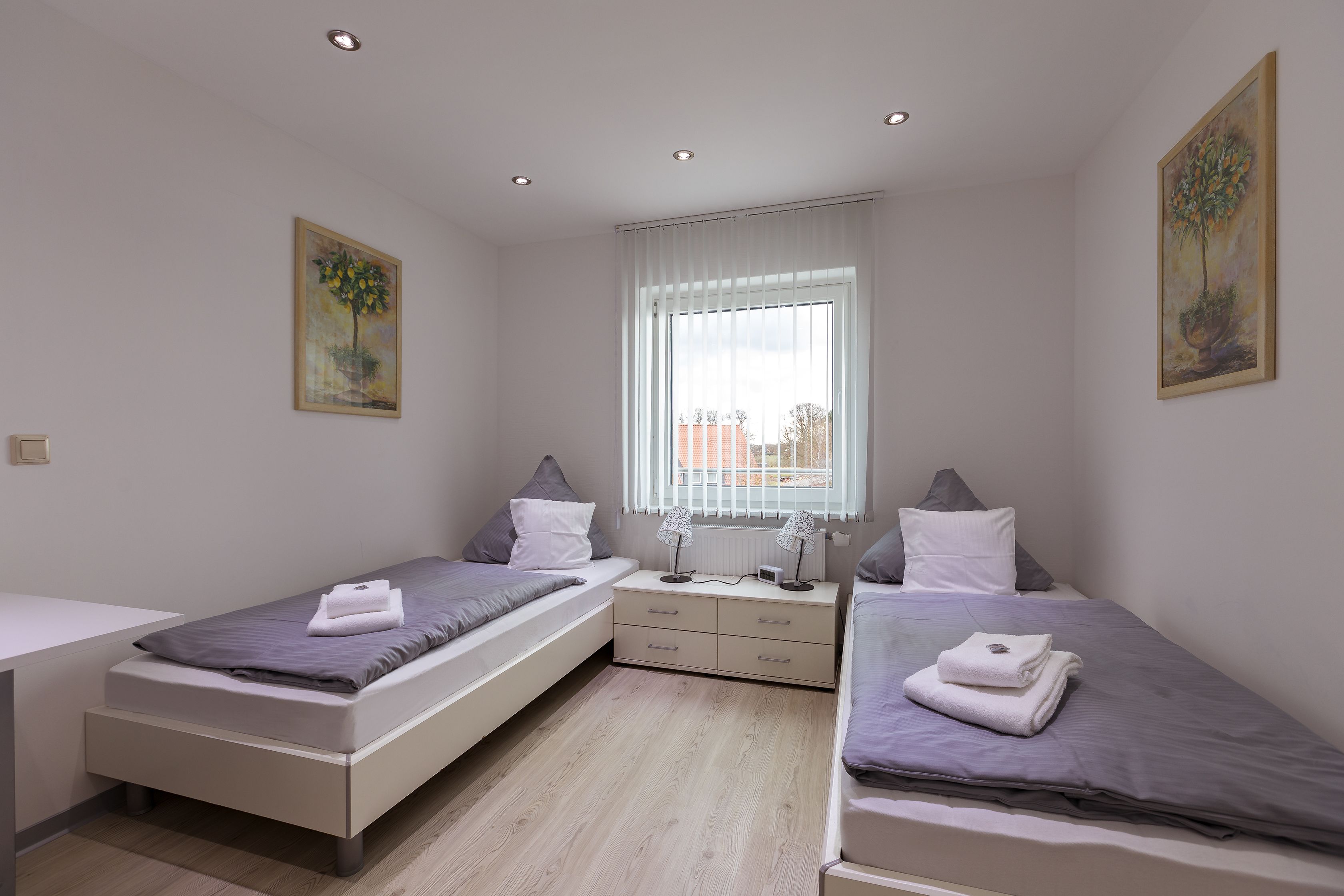 A modern bedroom with two single beds, a nightstand, a dresser, and a window with blinds.
