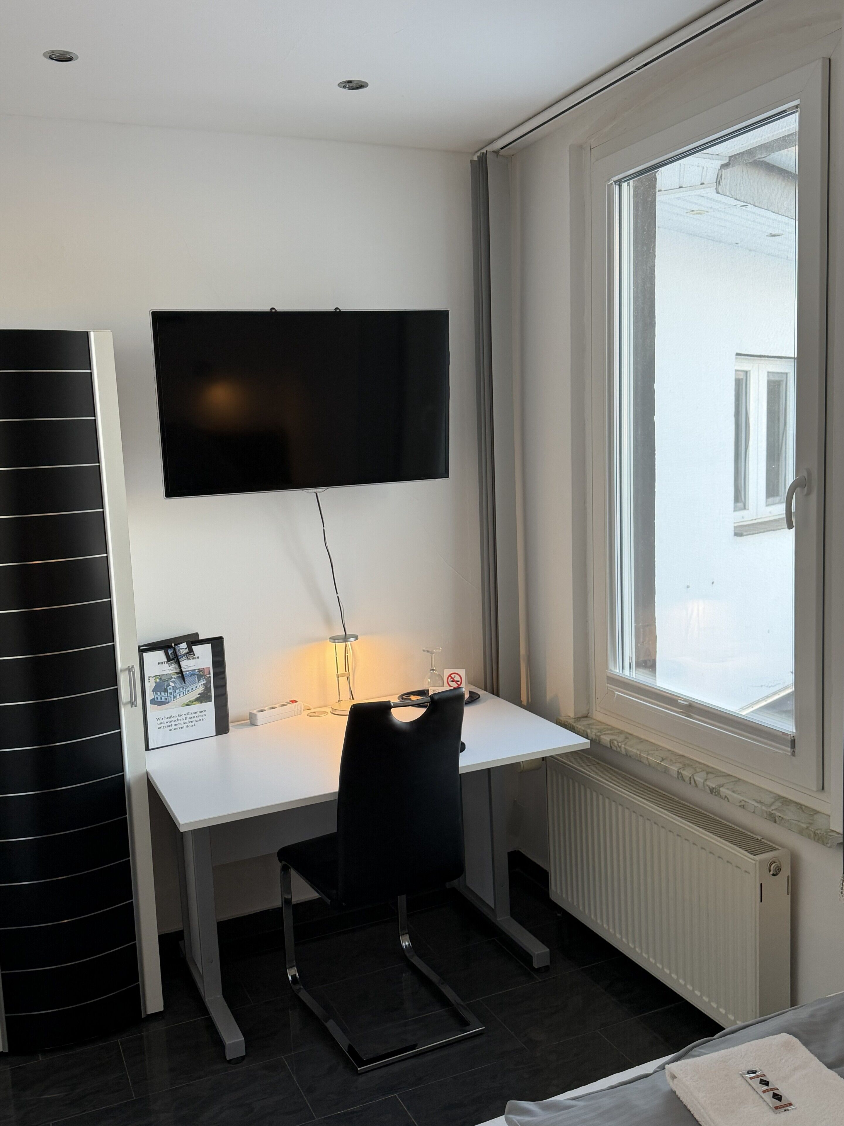 A hotel room with a desk, a flat-screen TV mounted above it, a window with a view, a radiator below the window, and a black chair.