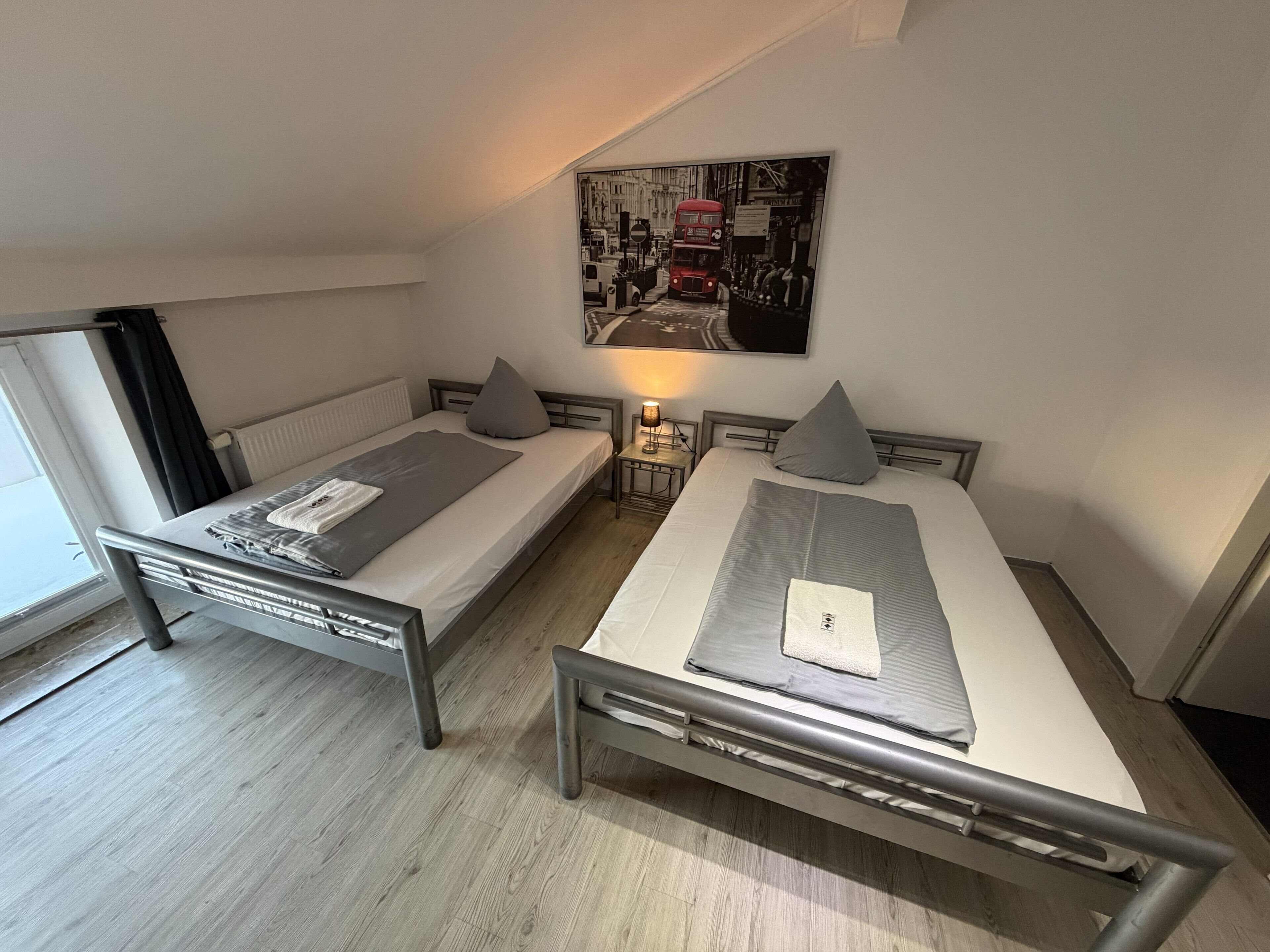 A twin room with two beds, a nightstand with a lamp, a radiator, and a cityscape painting on the wall.