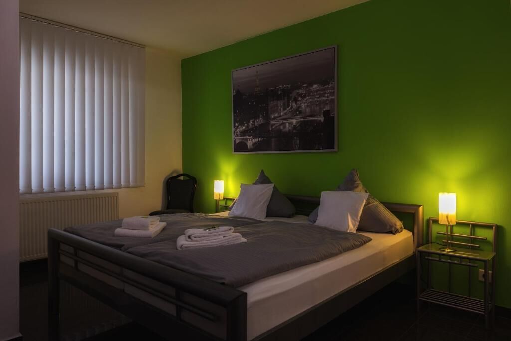 A hotel room with a bed, a nightstand, a lamp, a window with blinds, and a wall-mounted picture.