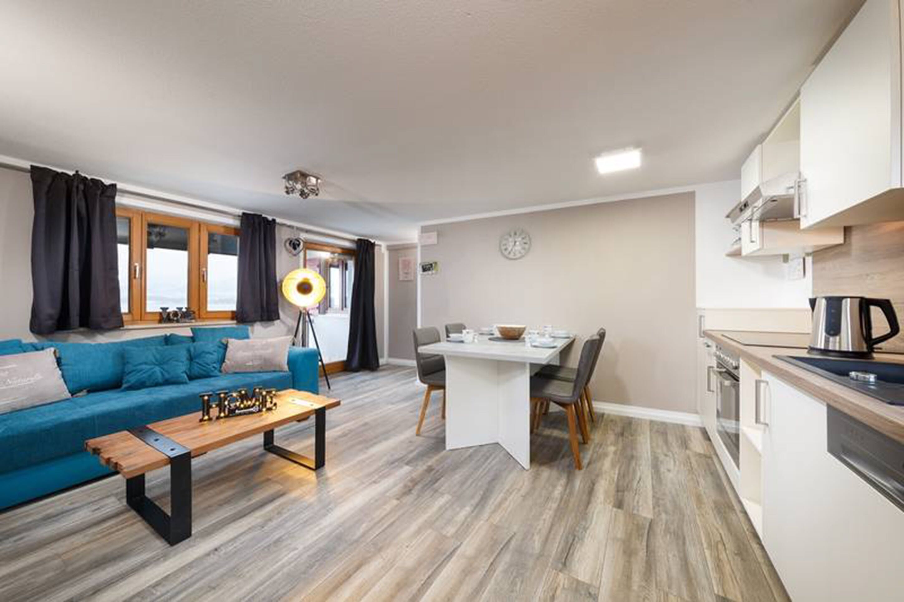 A modern living room with a sofa, coffee table, and dining table. There is a kitchen area with cabinets and appliances.