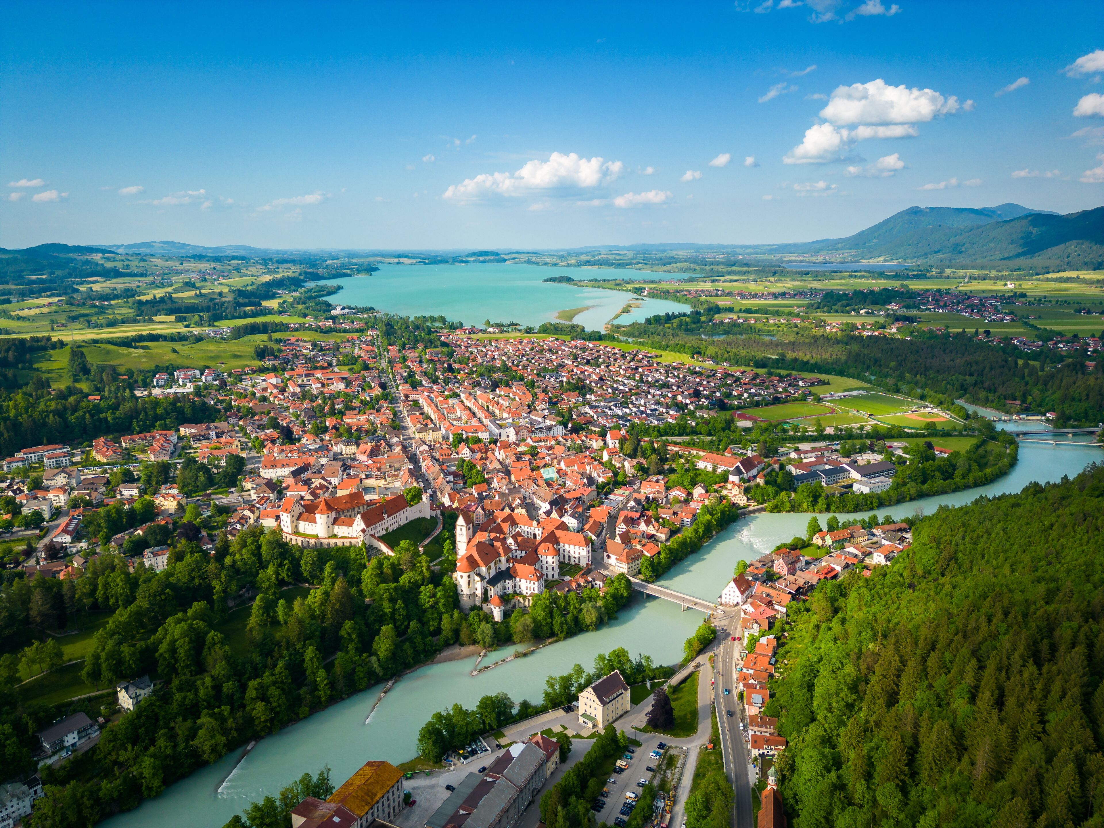 An aerial view of a town with a river, surrounded by greenery and mountains.
